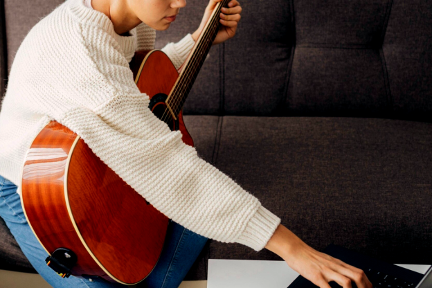Moça sentada no sofá, diante do notebook, com o violão no colo, aprendendo música online